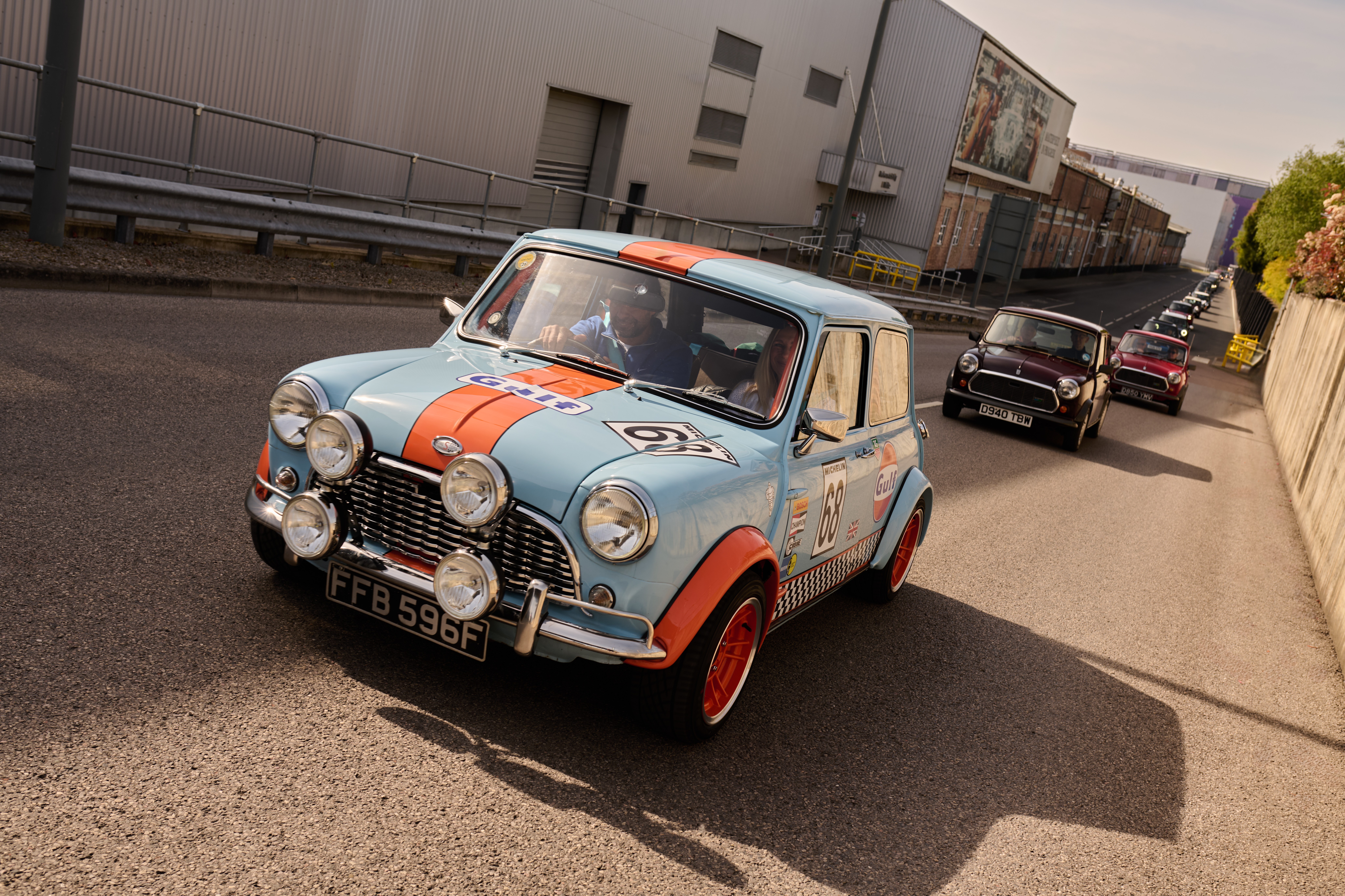 People gather outside a large black building, admiring a row of classic and modern Mini cars.
